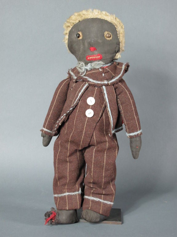 circa 1920<br />
This doll is wearing a chocolate pinstripe suit with light<br />
blue yarn details on edges. His face is primitively <br />
embroidered with small black button eyes. <br />
His hair is made of a small remnant of white fur.