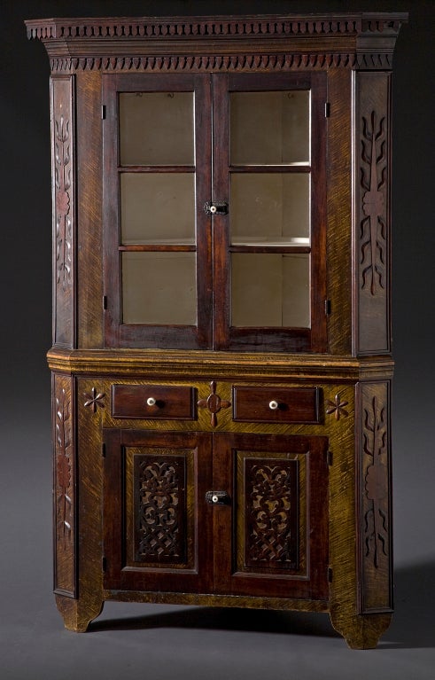 Pine 
Elegant corner cupboard with faux paint details of 
mustard and cherry throughout. Applied wooden floral 
motifs trim corners and center doors. Two Piece. 
Three pane glass doors with porcelain knobs. 
Ornate fence post cornice. High bracket