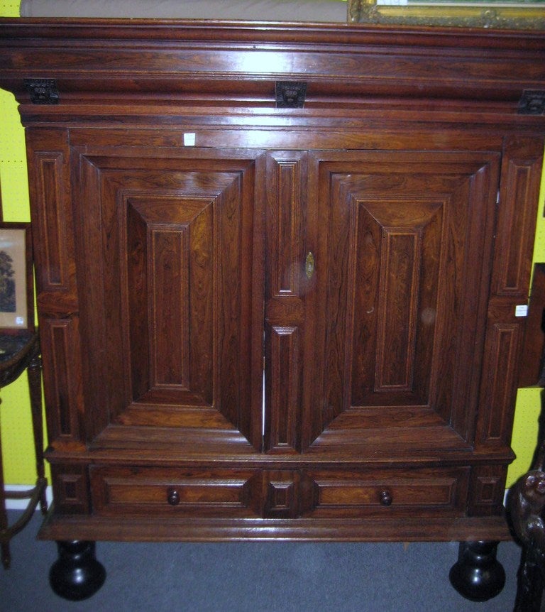 Dutch carved oak and parcel-eboninzed cushion cabinet, With a detachable molded cornice with carved masks above a pair of cushion paneled doors over a base with two drawers on two bulbous ebonized feet.