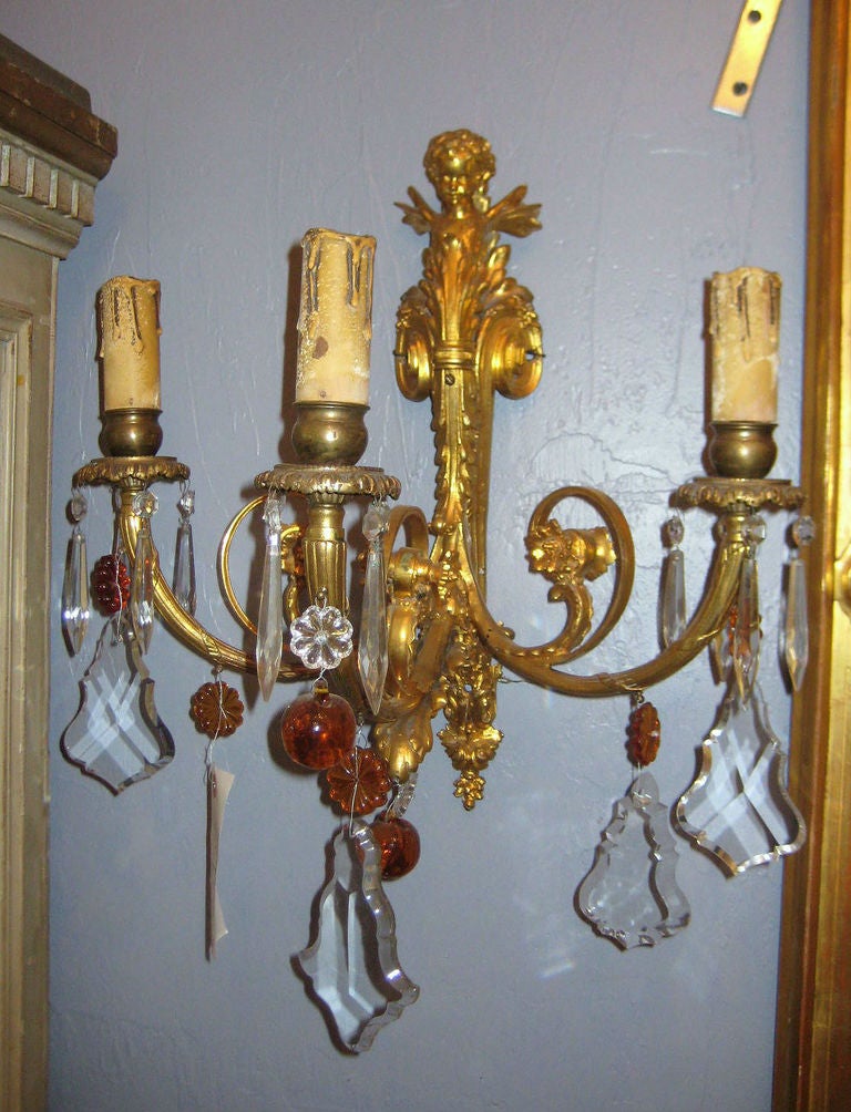 A fine pair of French gilt-bronze three-light wall sconces, each having a figural back plate, scroll and foliate design and issuing three candle branches with bell-form candle cups and floral bobesches suspending colorless crystal beads, spikes and