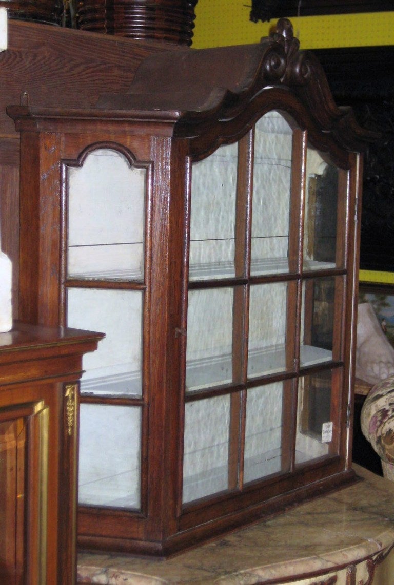 19th century Dutch carved oak hanging cabinet, having a molded arched cornice centering a carved fleur dis lis (also spelled: fleur de lys, fleur de lis) above an arched door with six original glass panes, the sides also with three original glass