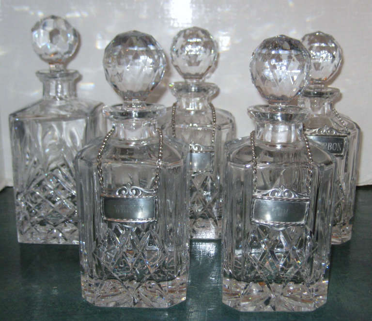 Five heavy crystal decanters and stoppers with pewter liquor labels, four square with sloping shoulders and one completely square; the four matching decanters with pewter labels for Brandy, Whisky, Rum and Bourbon; all with Baccarat style star