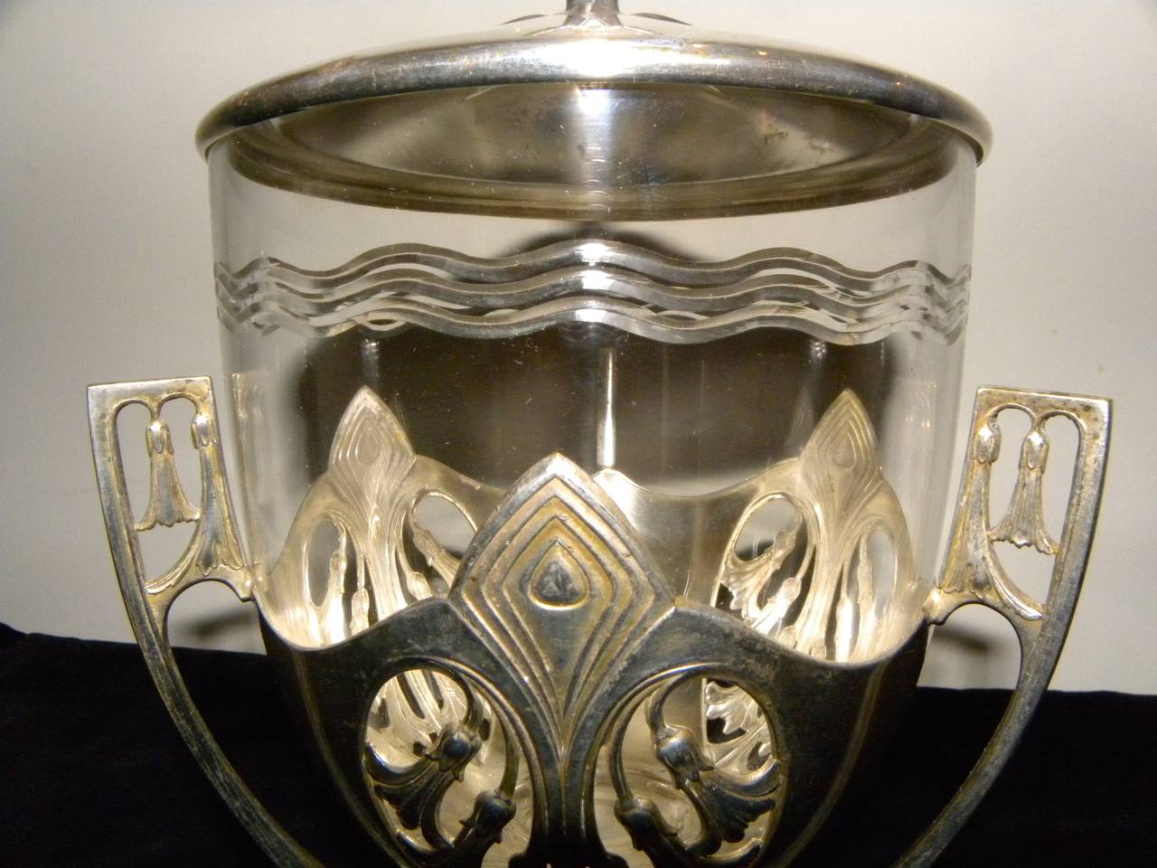 An etched glass urn with an intricate Art Nouveau silver stand and lid has all the Jugendstil elements that are the Harbinger of “Art Deco” that would develop over the decades that followed its creation in 1910. It is ornamental, but with clean