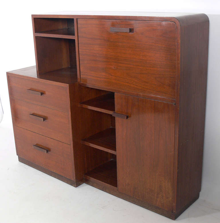 Art Deco Asymmetrical Bookcase / Credenza / Drop Front Desk by Modernage, American, circa 1930's. It offers a voluminous amount of storage with numerous drawers, shelves and compartments, and opens to reveal a drop front desk with articulated lamp.