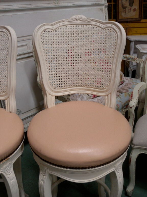 A matching pair of stunning bar stools, vintage to 150-60's.  French cane backs, scalloped aprons, carved legs, center metal foot rest and carved crest, all in a Cottage white paint.  Original pink naugahyde seats that SWIVEL!