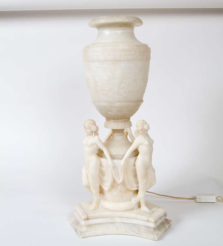 Carved white alabaster neoclassical style urn featuring three carved graces and carved classical attributes. Urn is illuminated by light bulb.