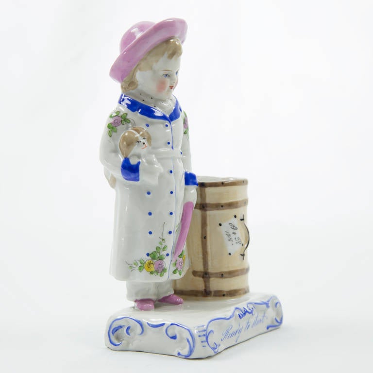 German porcelain spill vase modeled as a young girl holding her Cavalier King Charles Spaniel in one hand and her pink umbrella in the other, standing beside her travel trunk. Raised on scrolling base with title 