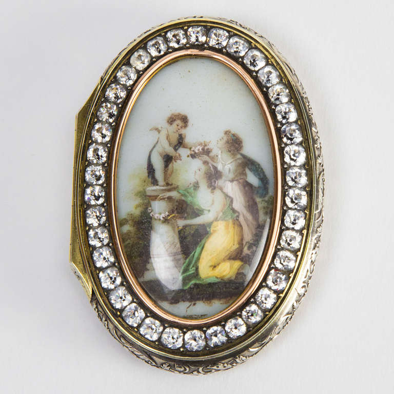 A superb antique silver gilt box, featured on the lid is a Porcelain image; beautifully hand decorated showing two Ladies showering a Cupid with flowers, surrounded by Paste stones. Outer edge wonderfully executed with detailed repoussé designs.