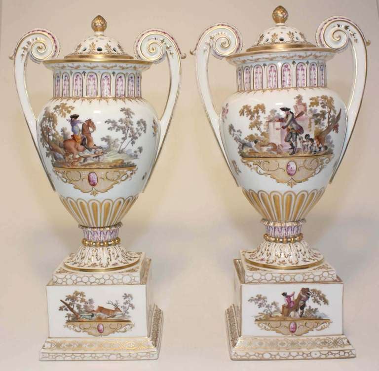 Pair of Dresden ivory porcelain parcel gilt footed potpourri vases with scrolling handles, hand-painted with individual hunting scenes, each centered by miniature city scene in monochrome puce, raised on similarly decorated square stepped bases.