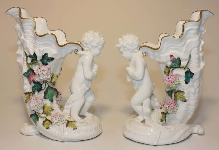 Pair of English figural porcelain vases, each modelled as a cherub pulling a scalloped cornucopia with applied polychrome flowers and vines.. 
They are not marked but are very much in the style of Moore Brothers