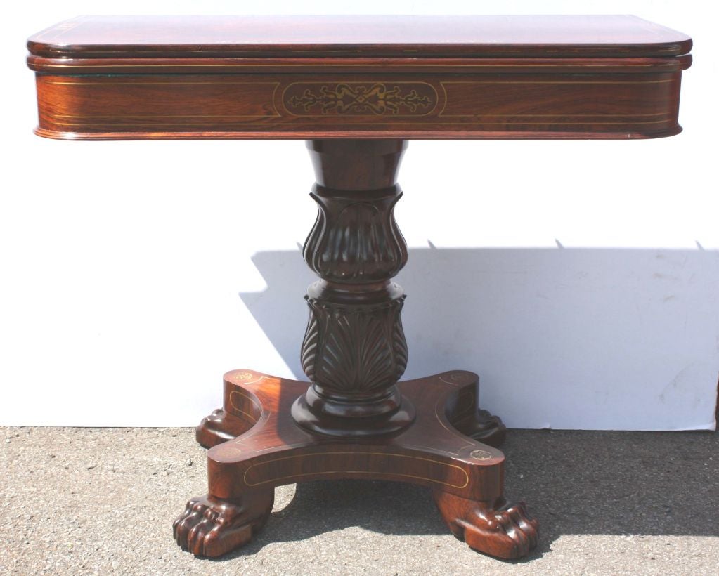 William IV strongly figured mahogany  brass inlaid fold-over games table, the shaped rectangular top with green baize and storage compartment, raised on a boldly carved pedestal, and quatrafoil base and paw feet.