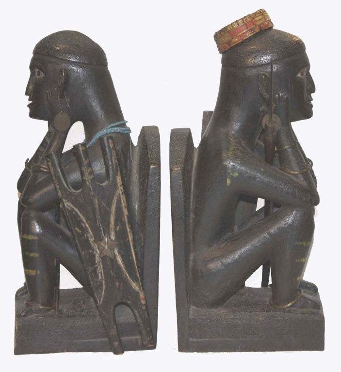 Pair of carved ebony bookends modelled as African warriors, resting on their haunches, with glass eyes, 19th century Spanish coin-earrings, brass bracelets, each with shield and spear.