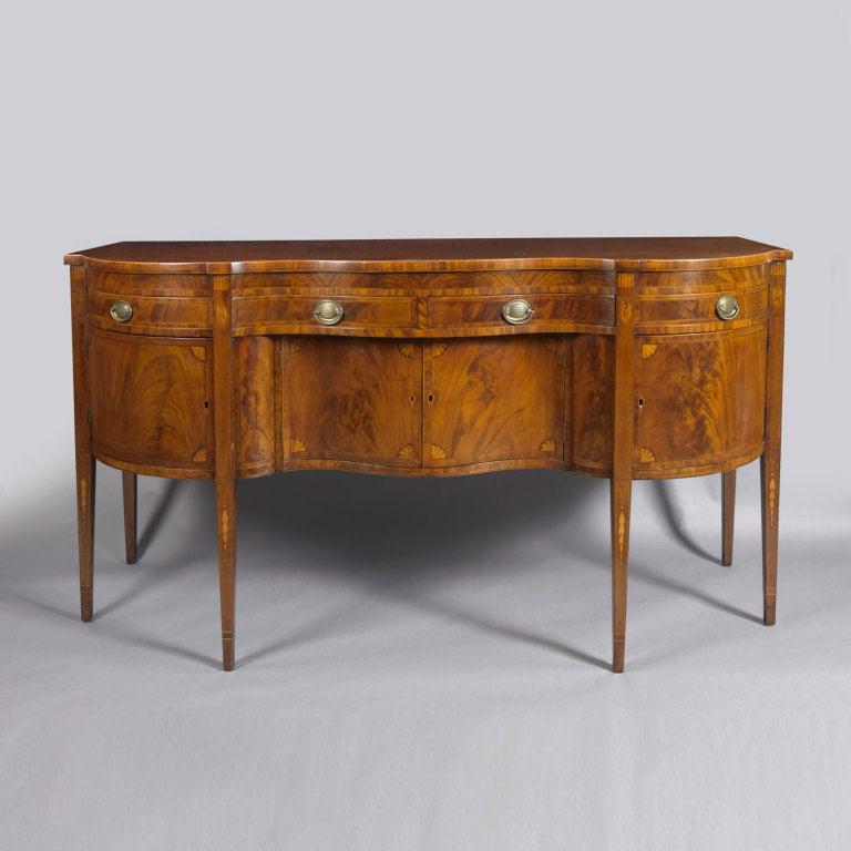 Probably New York, circa 1795-1810.
The form of this sideboard is typically found in New York. The serpentine front single board mahogany top conforms to the centre of the case which has two drawers above two doors which are inset. The ovolo sides