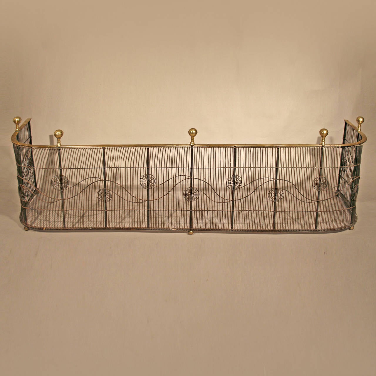 Late Federal wirework fender.
American, circa 1820.
Brass, rounded posts, forged iron, wirework.

This fully developed fender is adorned with rounded brass finials above a brass rail above intricate wire work with scroll motifs supported by