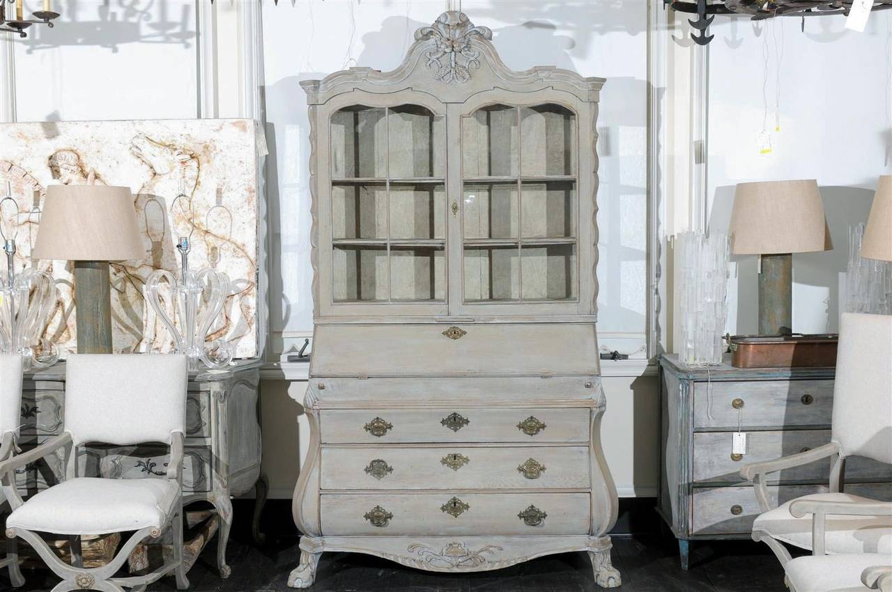 A European 19th century glass door cabinet with three lower drawers and drop front desk. The European secretary has multiple drawers in the desk part with fake books sections. It also features a bonnet with a nicely carved crest. The glass doors