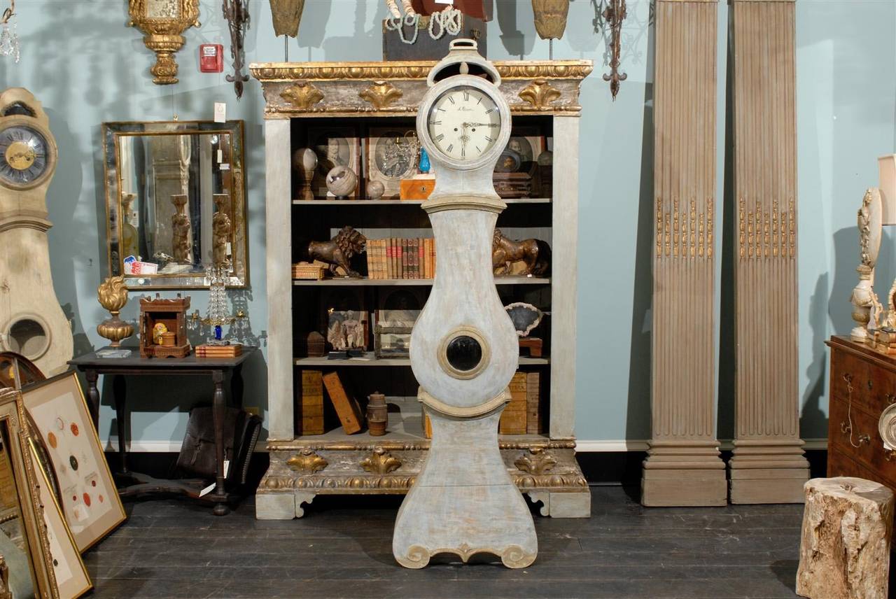 This painted Swedish clock stands on a base accented with a diamond shaped carving between the two lovely scroll feet.   

The middle section of this clock is fully fronted by a teardrop shaped door featuring a round convex glass window. 

The