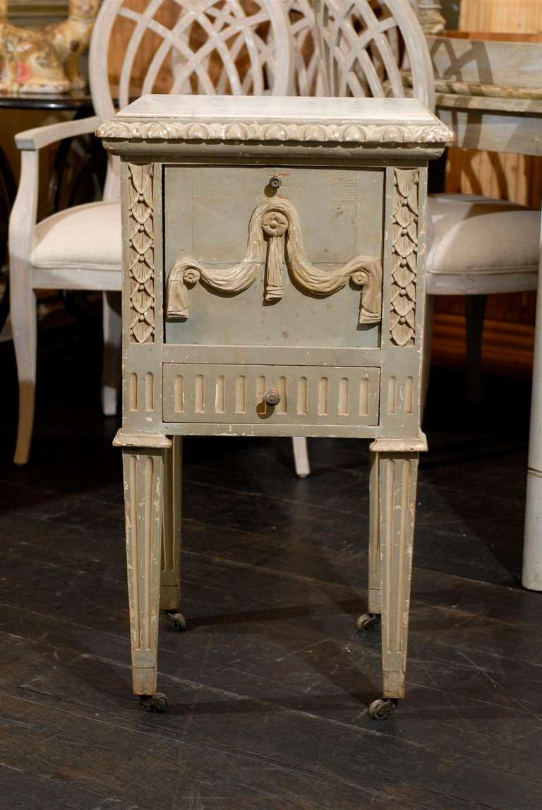 A French late 19th early 20th century richly carved wooden drop-front nightstand table / chest with white marble top, on casters with one lower drawer and egg and dart molding around the top. This French nightstand table features its old paint made