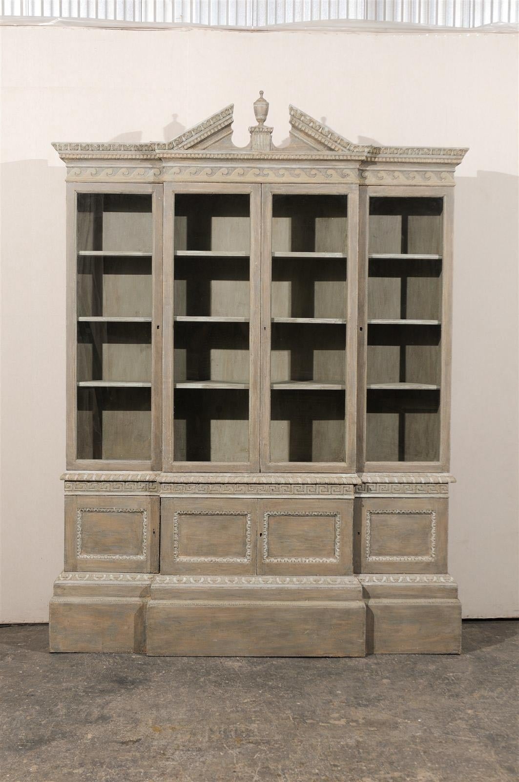 An English painted wood breakfront cabinet with glass doors, broken pediment with carved urn and various carved friezes, including Greek key and dentil molding, 20th century.