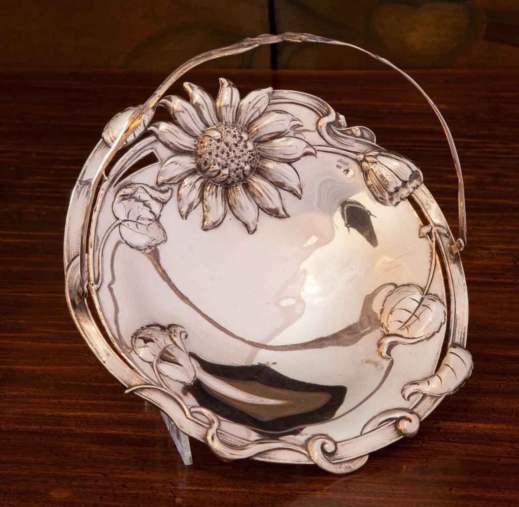The circular dish having openwork sides formed by leaves and branches, one rim decorated with a large relief sunflower, an open daisy, a closed flower and entwined leaves and branches; mounted with a slender bail handle with a chased flower and bud