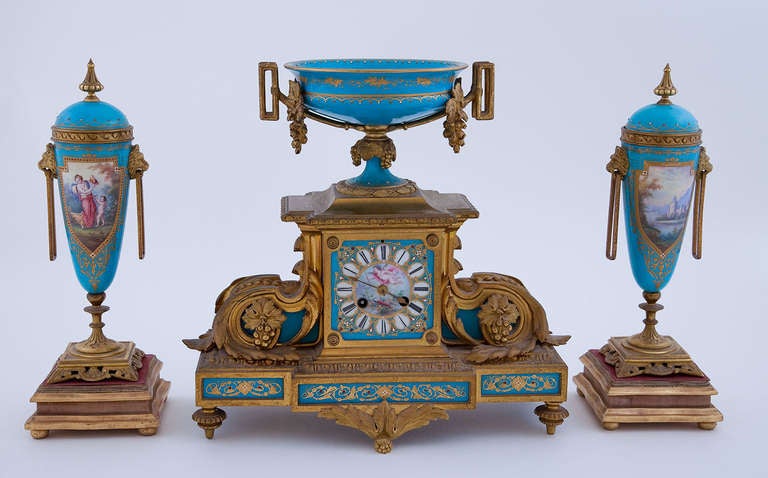 Comprising a pendulum clock with tazza, & two covered urns in bleu celeste.

The clock of architectural form the enamel painted dial sporting a flying cupid with arrows and lyre flanked by gilded volutes in the form of acanthus leaves with grapes