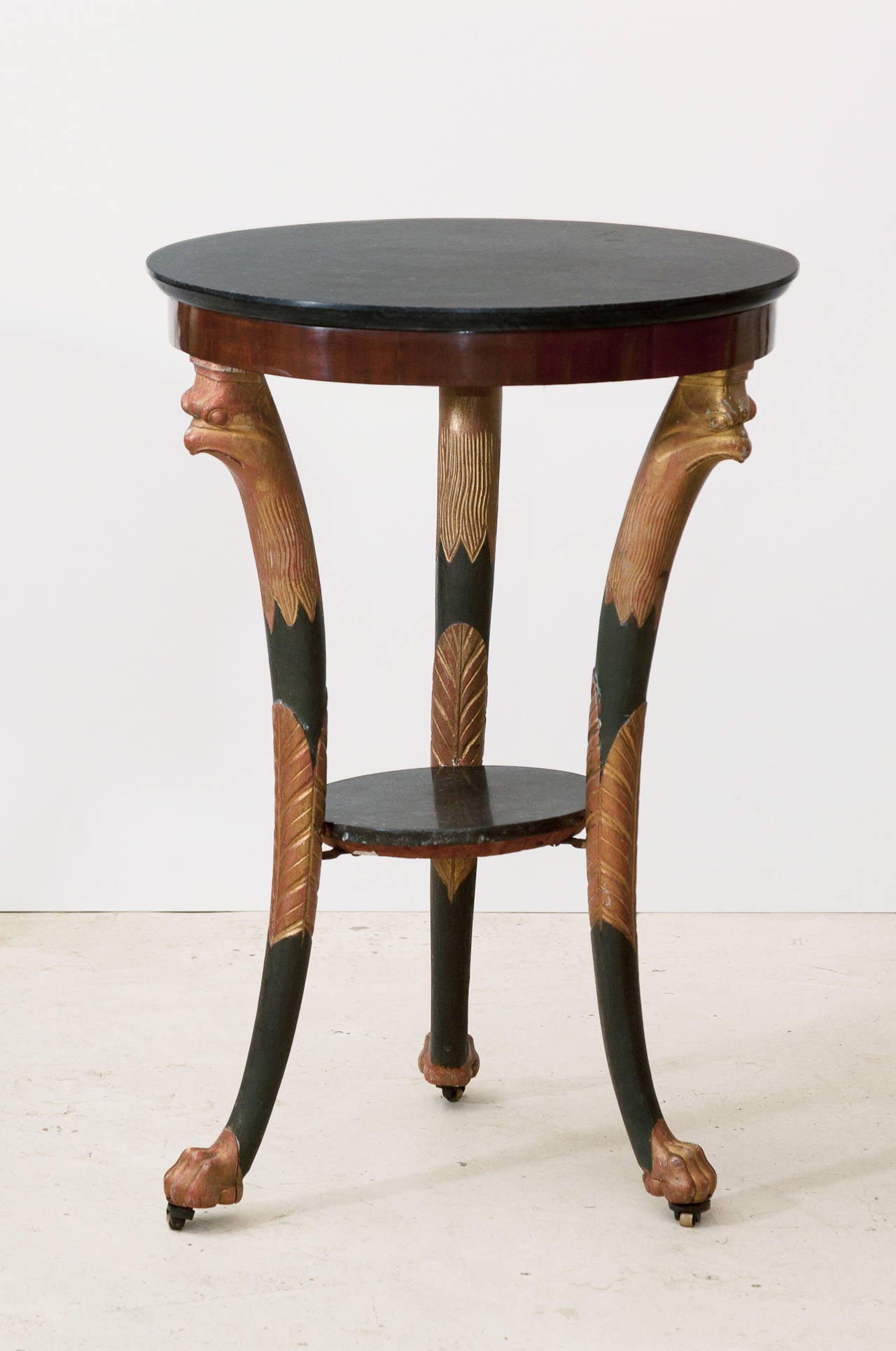 Standing on three legs partly gilt and painted to resemble bronze. Matt black fossil marble top. The tops of the legs with carved falcon heads in the Egyptian taste, supports joined by a circular undertier with matt black fossil marble insert