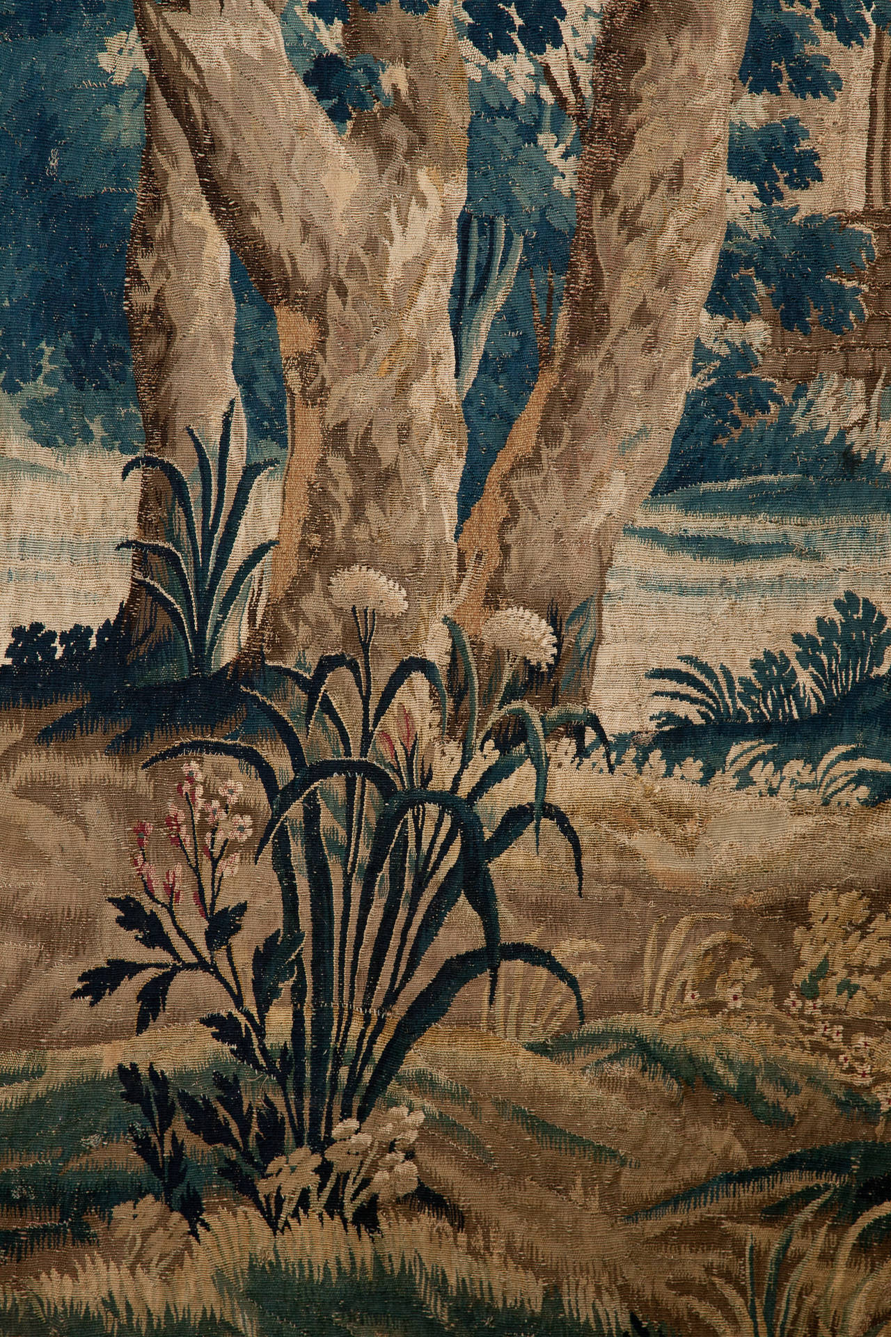 Large fine quality verdure tapestry with trees arching over to meet in the centre, a stream in the foreground has two small wading birds, behind is a bridge and a view of the doorway into a building. Surrounded by a rich border of interlaced fruit