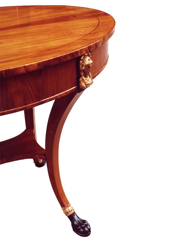 Superb oval Biedermeier side table in cherry on pine, inlaid with plum-wood. Apron with four fine carved Egyptian gilt-wood busts over four elegantly shaped saber legs, connected with a shelf and ending in ebonized paw feet with gilt-wood carvings.