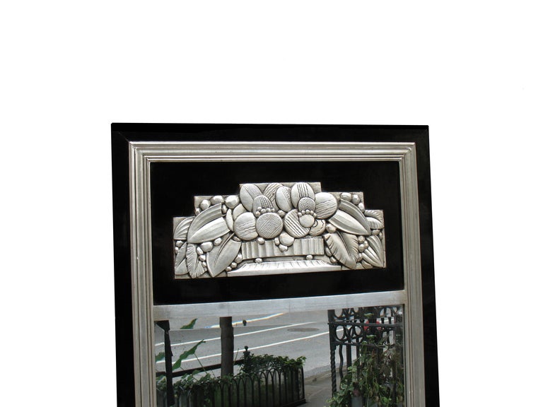 This design with fluted silver leaf moldings inside the black lacquered frame lengthens this pier mirror or wall mirror optically. The black panel with a wood carved silver leaf relief decoration depicts characteristically Art Deco motifs, like
