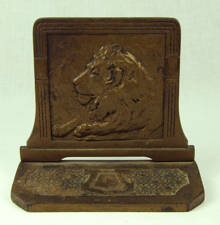 Featured is a set of brass bookends in a deep bronze color. A resting lion is carved in relief and framed with deco styling. The back of each bookend has an allover design and is stamped with 