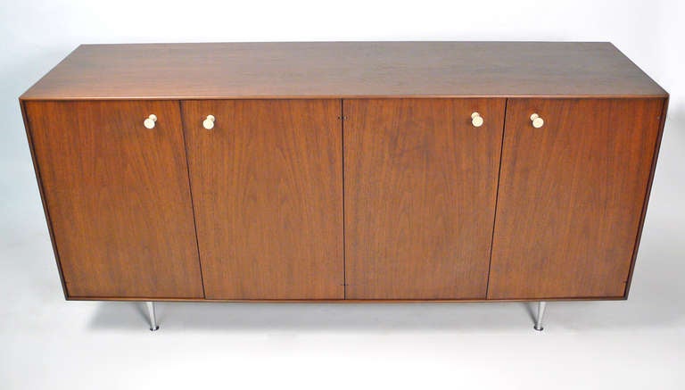 Mint condition walnut George Nelson thin edge credenza with porcelain hourglass pulls and original Herman Miller paper hanging tag. The cabinet consists of four hinged doors concealing adjustable shelves and stands on cast aluminum champagne glass