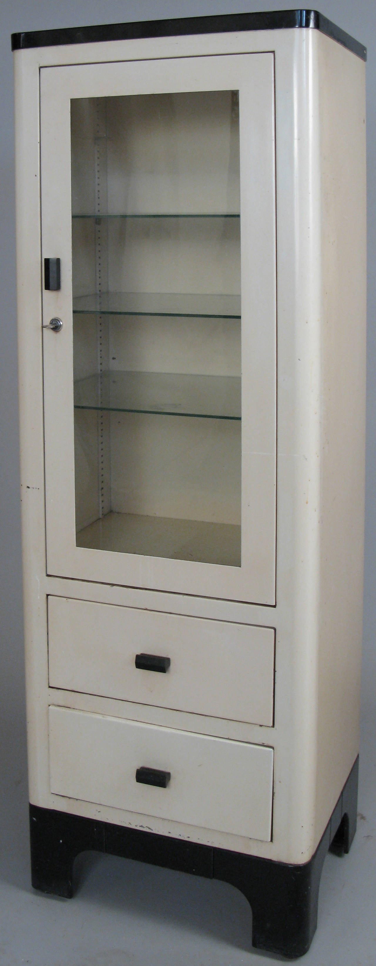Ein klassischer moderner cremefarbener Medizinschrank aus den 1940er Jahren mit abschließbarer Glastür:: verstellbaren Innenregalen und 2 unteren Schubladen. in seiner originalen cremefarbenen Ausführung:: mit schwarzem Sockel.