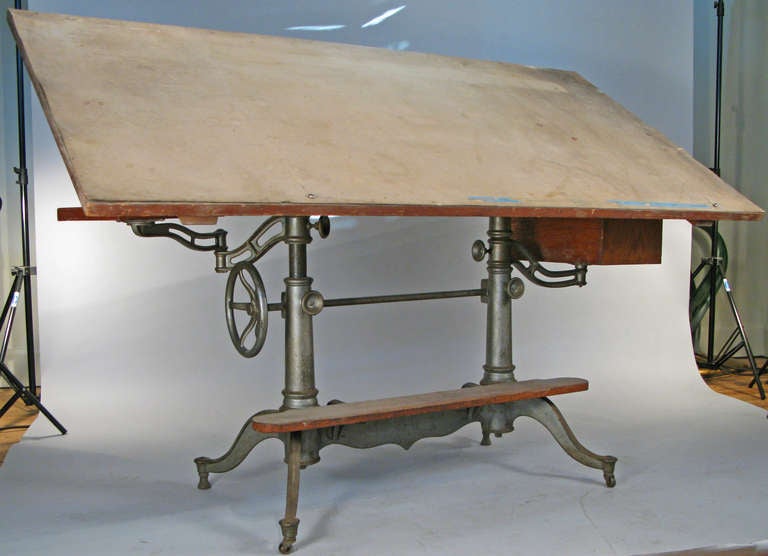 a very handsome antique 1920's fully adjustable drafting table by Dietzgen, with a cast iron base and adjustable swing arms on both sides, one supporting a tray, and the other a case with 2 storage drawers. includes the original footrest with cast