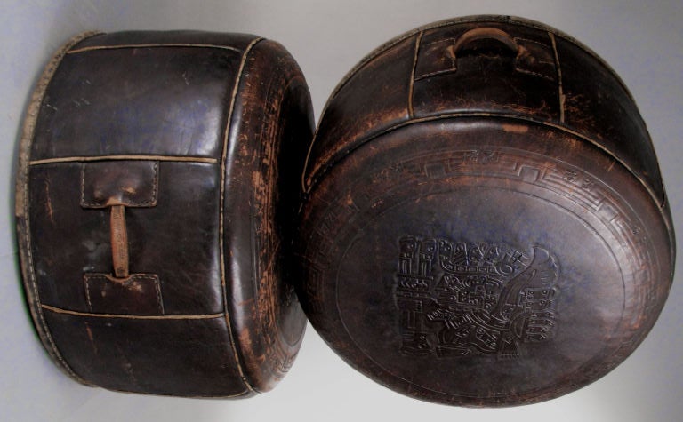 a beautiful pair of antique round hassock ottomans in thick saddle leather. beautifully made, each with a leather handle. the top of each hassock is tooled with a landscape filled with animal and nature scenes. the leather has a beautiful patina.