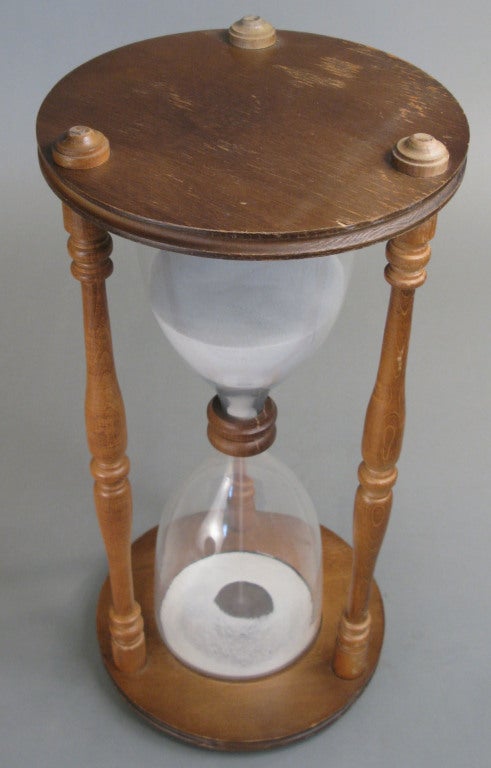 a very large antique blown glass hourglass, with sand running approximately one hour. encased in a wood base and top with 3 turned wood spindles.