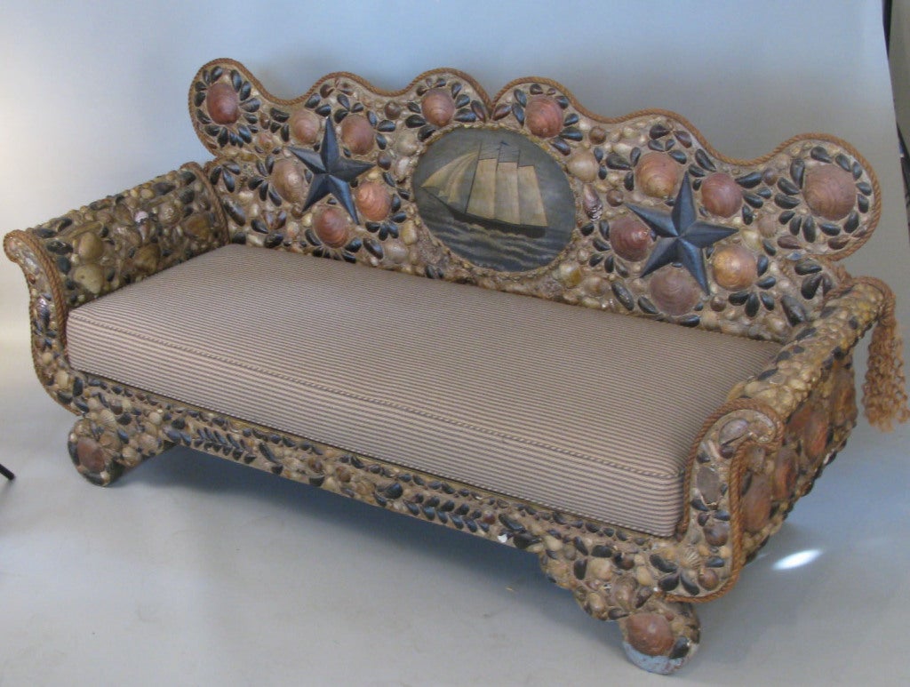 a wonderful nautical theme sofa with a gracefully curved frame encrusted entirely with patterns of natural seashells, with rope trim and a centerpiece of an oil on board painting of an antique schooner. seat upholstered in ticking.