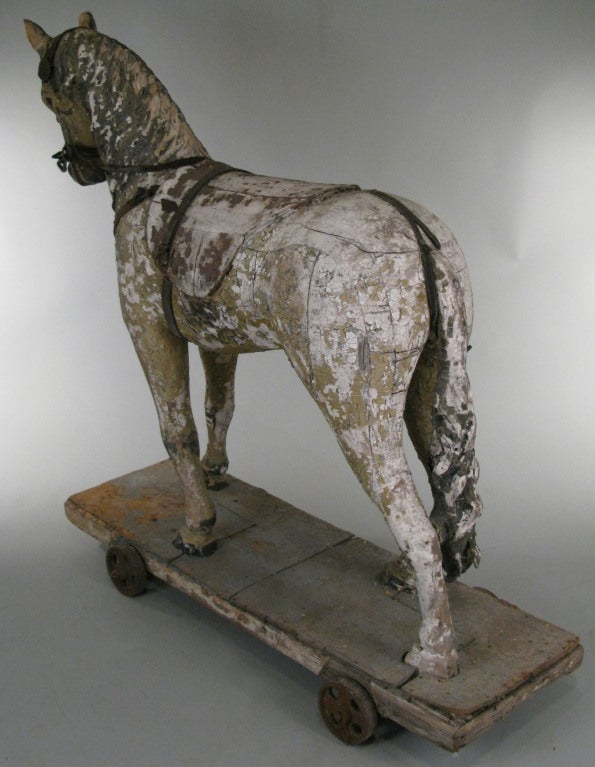 an anitique half-life size carved and paint decorated wooden horse, mounted as a pull toy on a wood cart with cast iron wheels. finish worn and cracked, with some parts of the leather harness remaining.