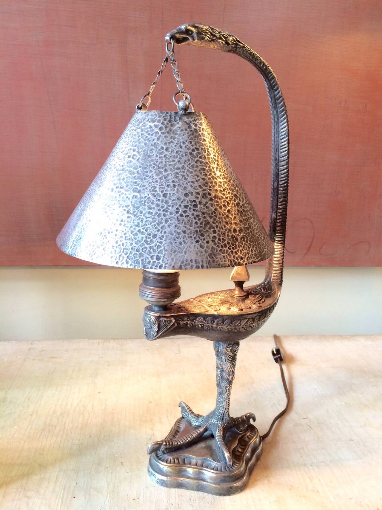 A rare neoclassic silver over brass whale oil form lamp mounted on eagle talon footed base with hand hammered shade suspended by chain from an eagle headed serpent form handle concealing a single Edison socket. Vintage electrical wire in excellent