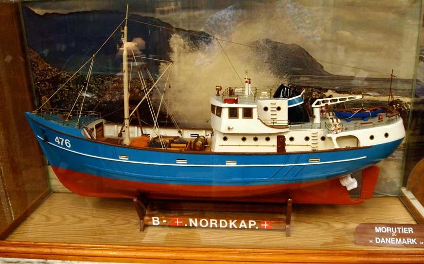 Painted ship model of 'B.Nordkap' enclosed in glass case with dramatic background of the coast.