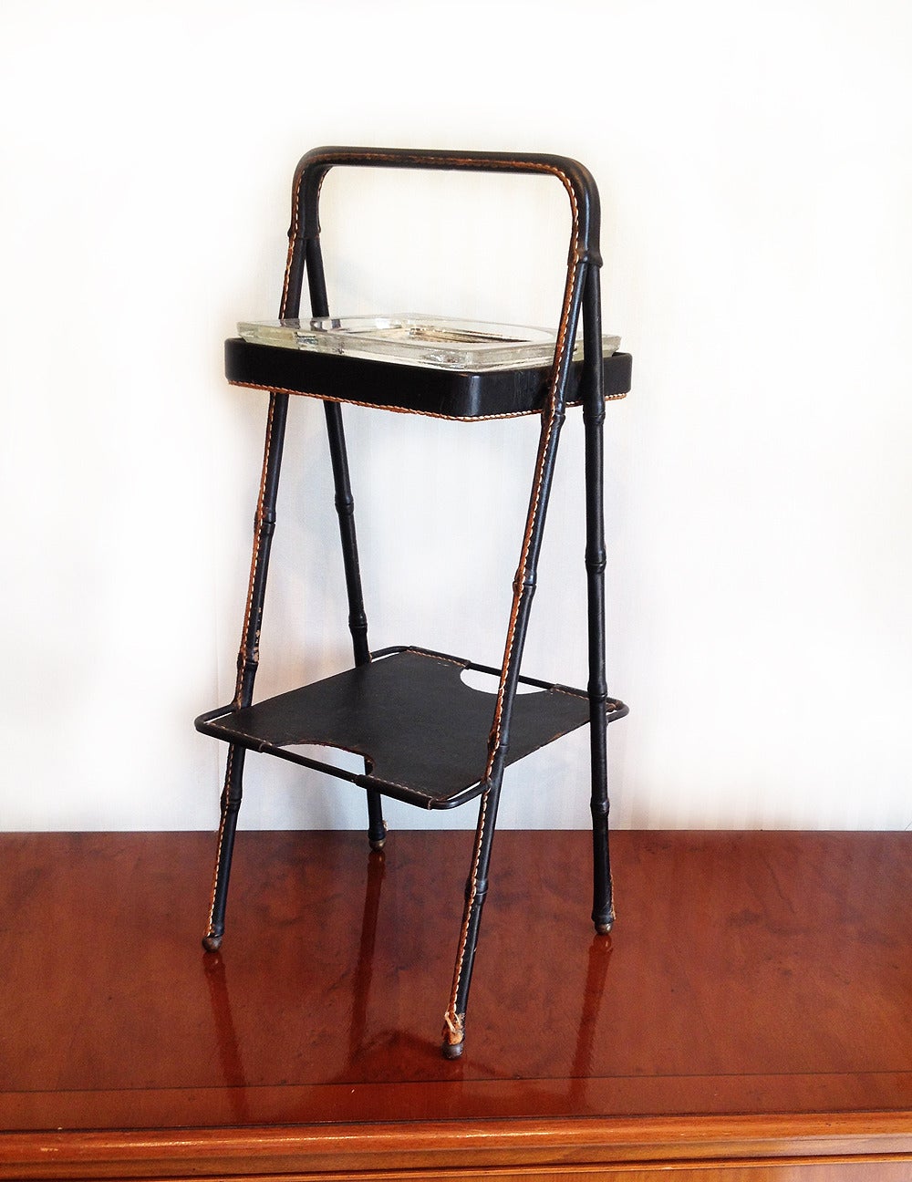 Beautiful smoking stand with stitched black leather frame terminated with four brass balls.
The glass ashtray or dish is in excellent condition.
Dimensions: H 23