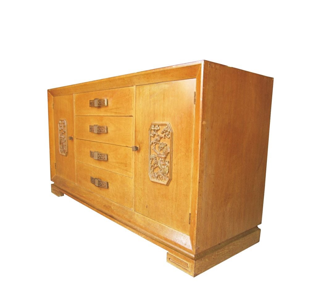 A bleached mahogany sideboard in the style of James Mont with carved wood plaques and brass handles. This piece has lots of storage room, it comes with four front drawers and it has one cabinet with a shelf and one lingerie drawer on each side. The