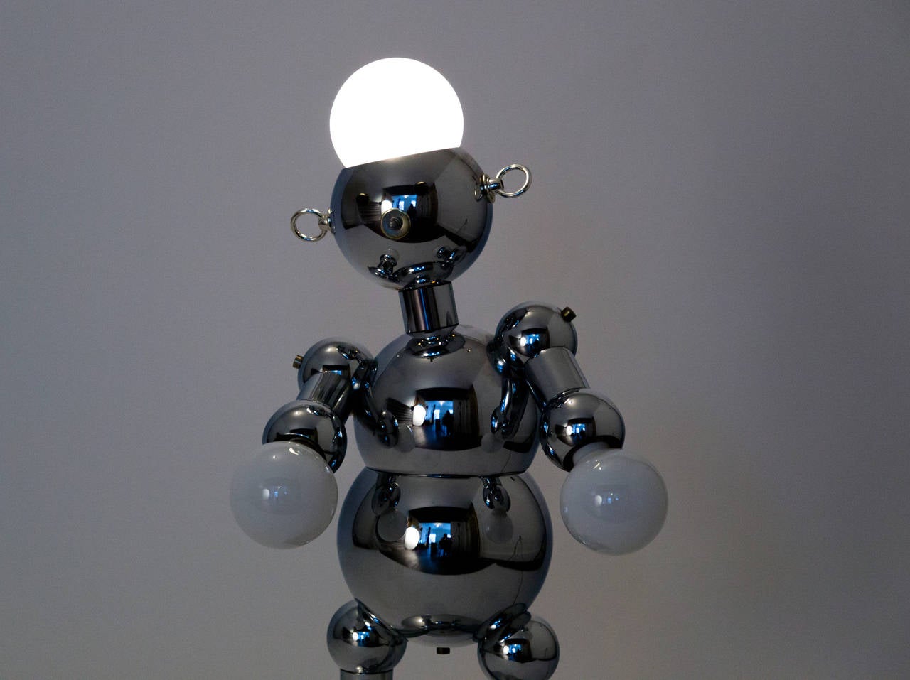 A charming, figural robot table lamp produced by Torino, comprising a segmented body in chrome, black resin eyes, metal ring fittings for ears, and a nose-switch that allows you to illuminate the head and the hands separately or together. We love