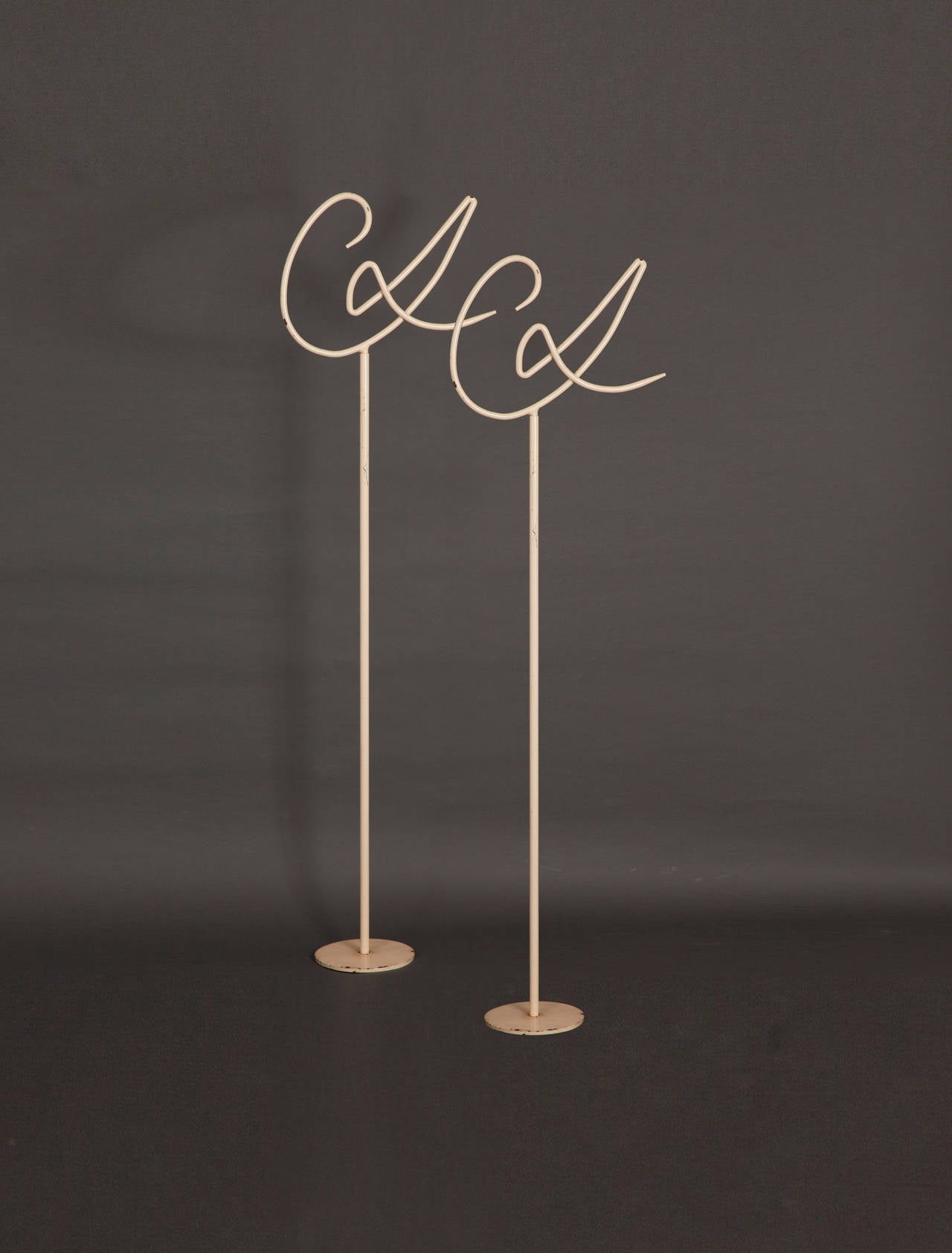 A pair of bent and enameled tubular steel display stands made expressly for fashion designer, Christian Lacroix, each comprising three individual components: a solid, round base, an upright, and a top sculpted as LaCroix's cursive 'CL' logo.

We