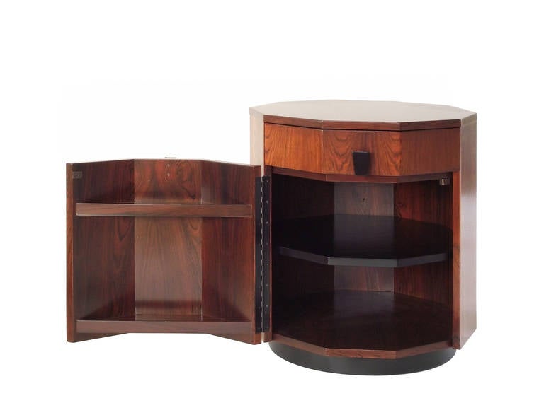 A dry bar or end table in a decagon form, executed in rosewood with ebonized, trapezoidal pulls and floating upon an ebonized plinth base, designed by Harvey Probber. The laminate-lined drawer and bottom compartment with adjustable shelf to the