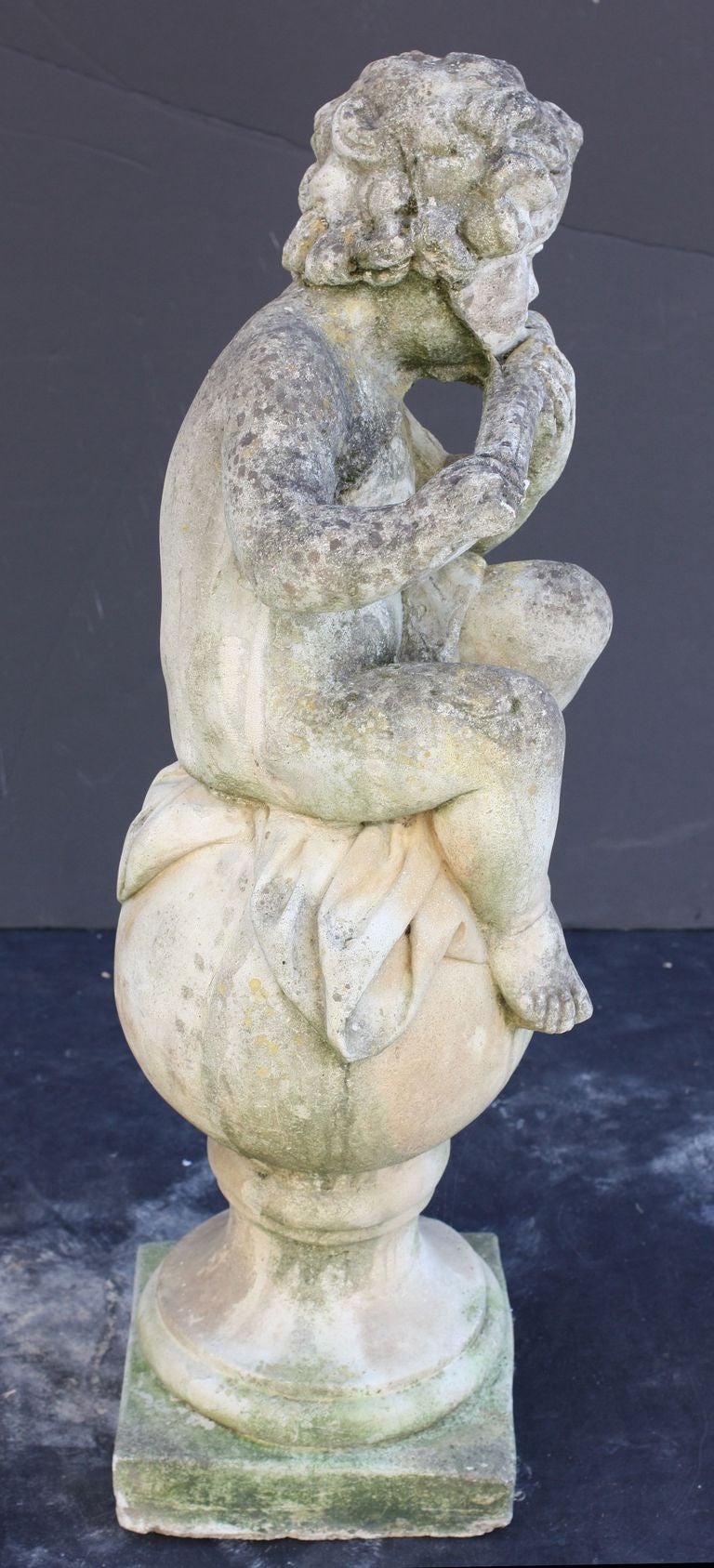 An English garden statue or ornamental figure of composition stone,   featuring a cherub or child putti in the Classical style, posed in a sitting position, playing a flute, on a ball and plinth base.
Great detail in the round.

Perfect for a