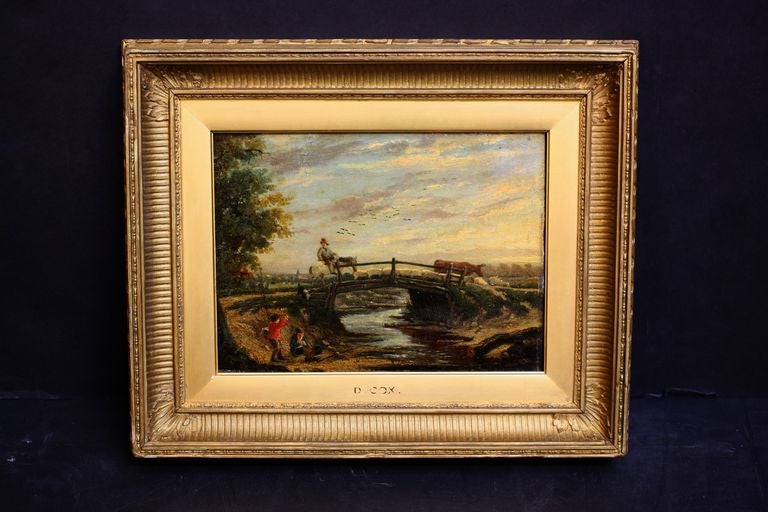 An English oil painting on panel, mounted to a gilt wood frame, featuring a rustic scene of boys fishing and a man herding animals over a bridge. 
Marked on frame: D. Cox