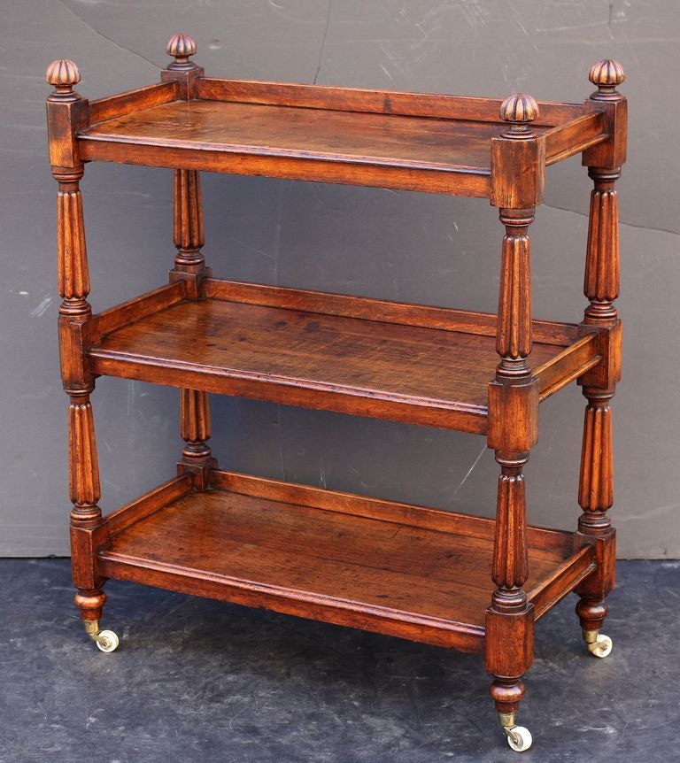 A trolley server (or dumb waiter) of oak featuring three tiers with edge-moulded tops, each with back gallery frieze, each tier adjoined to four turned column supports topped with finials, set upon rolling brass casters.

Such servers (often