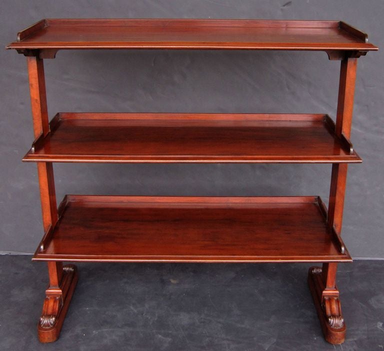 A trolley server (or dumb waiter) of mahogany featuring three tiers with edge-moulded tops, each panel with back gallery frieze, each tier adjoined to two turned column supports, with decorative acanthus leaf and scroll accents, set upon rolling