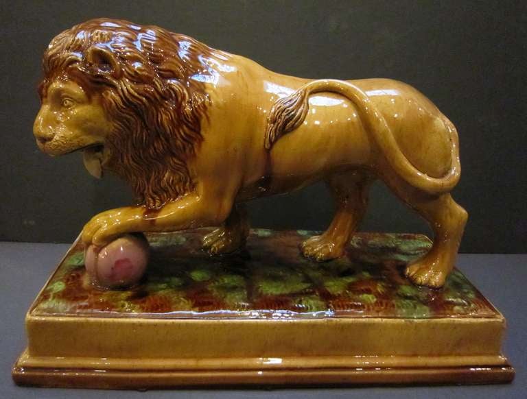 A handsomely modeled English lion with treacle glaze featuring a standing lion with left front leg on a ball and mounted on a rectangular plinth, from the Staffordshire Potteries.