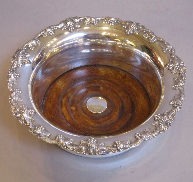 An English wine or champagne bottle coaster (holder) featuring a decorative rolled-edge top with a design of grapes and grape leaves, and an oak interior base with a silver cartouche.
Can be used as a soda siphon holder for the cocktail