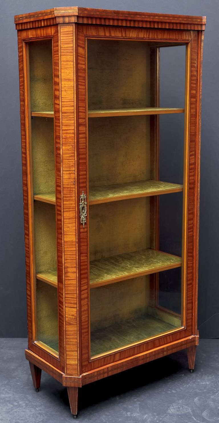 A French display case or china cabinet (or bookcase) of satin wood, featuring a canted front with choice veneers of satin wood, door with gilt ormolu escutcheon (and key), opening to three fitted shelves, and standing on tapered legs.